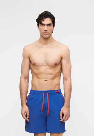 Νέος άνδρας με σκούρα σγουρά μαλλιά, χωρίς πουκάμισο, φοράει μπλε μαγιό με κόκκινο κορδόνι και ρίγες στα πλάγια, στέκεται μπροστά σε απλό φόντο.