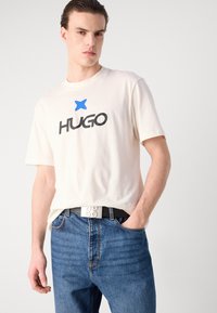 Beige cotton t-shirt with a blue star and black "HUGO" logo, paired with blue denim jeans and a black belt featuring a white "HUGO" buckle.
