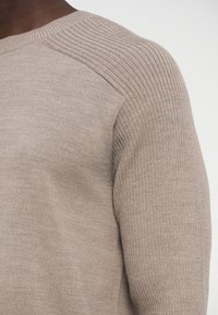 Beżowy prążkowany sweter z dzianiny z okrągłym dekoltem i fakturowanymi detalami na ramionach, pokazujący zbliżenie na tkaninę i szwy.