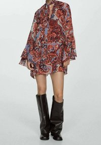 Langärmliges Kleid mit Paisleymuster in Rottönen und Blau, kombiniert mit schwarzledernen, knielangen Stiefeln. Fließender, transparenter Stoff.