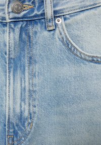 Gros plan sur un jean en denim bleu clair montrant une poche avant, une passants de ceinture, un bouton en métal et un rivet avec des détails de couture visibles.