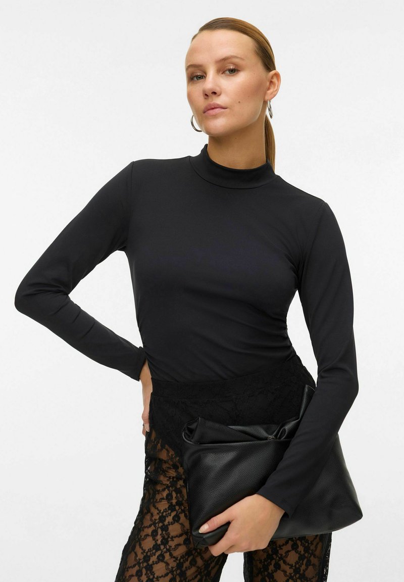 Černý dlouhý rukáv top s vysokým límcem, v kombinaci s nohavicemi zdobenými krajkou. Modelka drží texturovanou černou psaníčko.