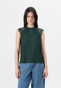 La modelo lleva una blusa sin mangas de color verde oscuro con encaje en los hombros, combinada con jeans azules, frente a un fondo blanco liso.