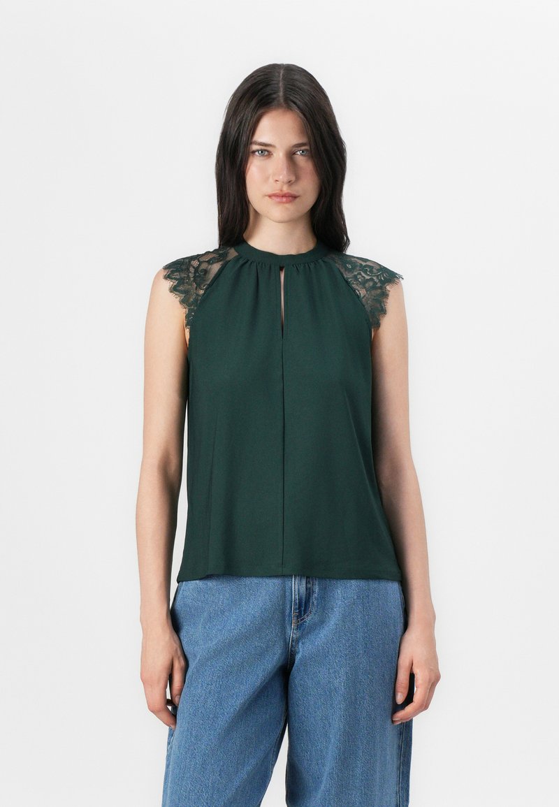 La modelo lleva una blusa sin mangas de color verde oscuro con encaje en los hombros, combinada con jeans azules, frente a un fondo blanco liso.