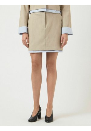 Ženska, oblečena v bež krilo s sukničastim kostimom s modrimi črtastimi manšetami in robom, črni čevlji z visoko peto, stoji ob enobarvnem ozadju.