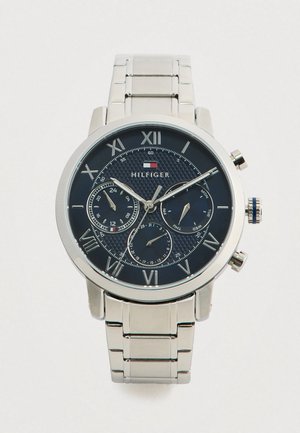 Reloj de pulsera de metal plateado con esfera azul, números romanos, tres subesferas para las 24 horas, el día y la fecha, y el logo de "Hilfiger" debajo de las 12 en punto.