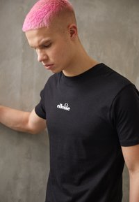 Black short-sleeve t-shirt with a white "ellesse" logo on the chest, made from cotton material. Model has short pink hair and leans against a wall.