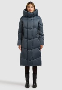 Párnázott, hosszú sötétszürke kabát magas gallérral. Hímzett mintával, két oldalzsebbel és egyenes szabással rendelkezik. Fekete leggings és csizma fölött viselve.