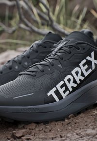 Scarpe da trail running Terrex nere con suole spesse su una superficie rocciosa all'aperto, con tomaia in mesh e logo bianco ben visibile.
