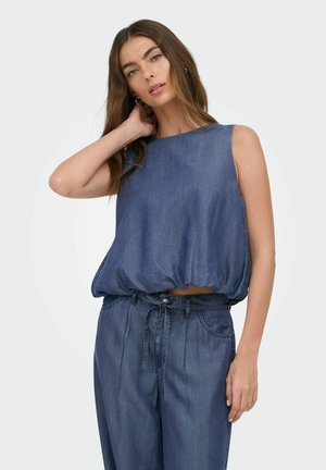 Femme aux longs cheveux bruns porte un haut ample sans manches en denim et un pantalon taille haute en denim assorti avec une ceinture nouée, posant avec une main derrière la tête.
