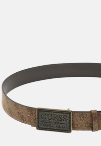 Ceinture en tissu marron avec le logo GUESS, comportant une section tissée à motifs et un dos en cuir noir lisse. Ajustable avec une boucle en métal.