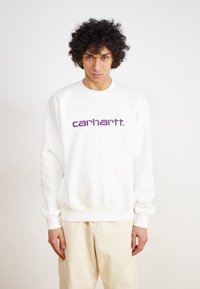 Sweatshirt blanc avec des poignets et un ourlet côtelés, arborant un logo "carhartt." violet bien visible sur la poitrine. Texture de tissu lisse.