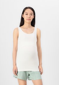 Λευκή μπλούζα εγκυμοσύνης με στρογγυλή λαιμόκοψη, ελαστικό ύφασμα και ραβδώσεις πάνω από την κοιλιά, συνδυασμένη με μint πράσινα σορτς.