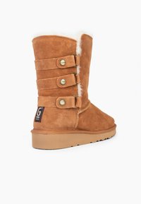 Bota de ante marrón con forro de piel sintética blanca, tres correas decorativas con botones dorados y suela de goma texturizada.