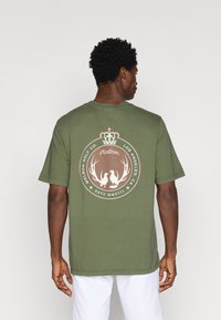 Oliņzaļš kokvilnas T-krekls ar apļveida logotipu brūnā krāsā, kurā attēlota kroņa un raga simbolika, kā arī teksts. Īsas piedurknes un brīvs griezums.