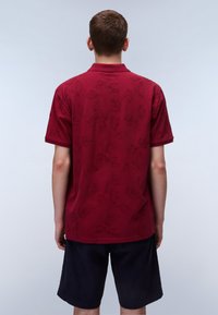 Burgundy pikétröja med bladmönster, korta ärmar och krage. Matchas med mörka shorts som visar en avslappnad passform och slät textur.