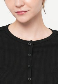 Črna rebrasta pletena majica z okroglim izrezom in šestimi črnimi gumbi spredaj, ki poudarja teksturirano tkanino in prilegajočo se zasnovo.