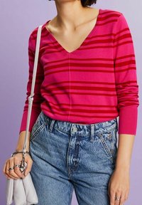 V-Ausschnitt Strickpullover in leuchtendem Pink mit roten horizontalen Streifen, kombiniert mit hoch taillierten blauen Jeans aus Denim und einer weißen Handtasche.