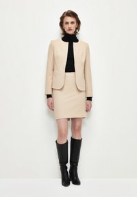 Blazer beige su un dolcevita nero, abbinato a una gonna a tubino beige e stivali neri al ginocchio. Texture liscia, design aderente.