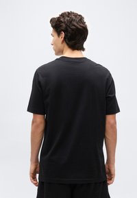 Άνδρας με σκούρα κυματιστά μαλλιά που φοράει απλό μαύρο κοντομάνικο μπλουζάκι, με την πλάτη γυρισμένη προς ένα απλό ανοιχτόχρωμο φόντο.