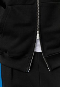 Sudadera con cremallera negra y puños y dobladillo acanalados, con una cremallera plateada y una capa interior blanca en contraste visible en la abertura.