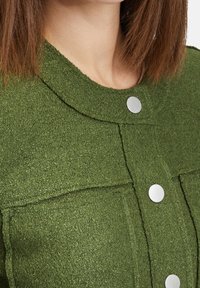 Green textured garment with a round collar, featuring silver snap buttons and visible stitching details.