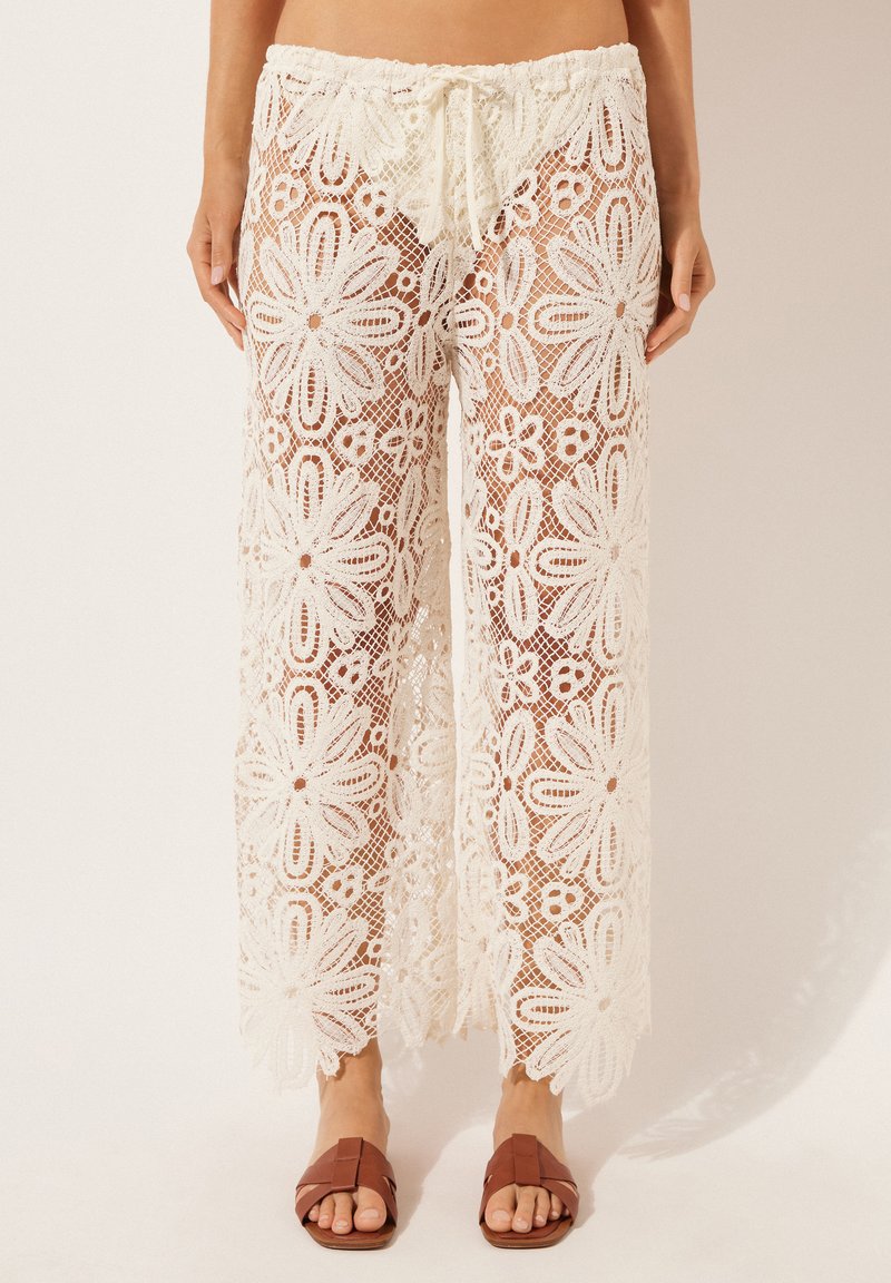 Pantalon en dentelle crochet blanc avec de grands motifs floraux, taille à cordon, porté avec des sandales plates marron à bout ouvert sur un fond neutre.