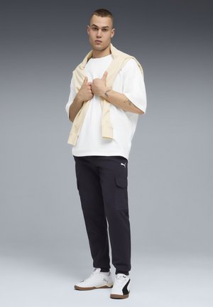 Joven con camiseta blanca, sudadera beige sobre los hombros, pantalones cargo negros de Puma y zapatillas blancas, de pie sobre un fondo gris.