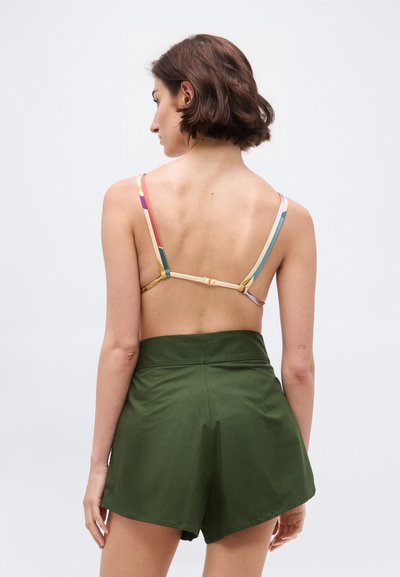 Kvinde med kort brunt hår iført en bikini top med regnbuestriber, der lukkes bagpå, og højtaljede grønne shorts, set bagfra.