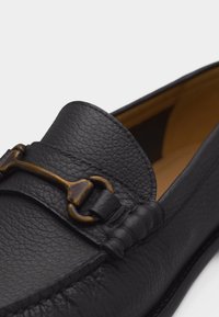 Μαύρο δερμάτινο μοκασίνι με υφή, με λεπτομέρεια από μπρούτζινο μεταλλικό κόμπο σε σχήμα αλόγου και ταμπά εσωτερική επένδυση, εμφανίζεται σε κοντινό πλάνο σε λευκό φόντο.