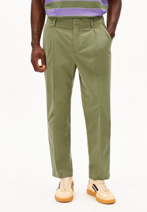 Mann trägt olivgrüne Hose, beige Sneakers mit schwarzen Streifen und ein lila-grün gestreiftes Hemd und steht mit Händen in den Taschen.