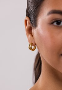 Goldene Creolen mit einem dreifachen Schleifen-Design, glatter Textur und glänzender Oberfläche, getragen am Ohr vor einem neutralen Hintergrund.