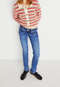 Cardigan a righe in rosso, beige e nero con grandi bottoni; jeans slim-fit blu; scarpe nere con una texture liscia.