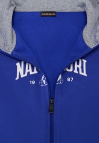 Sudadera azul con cremallera y cuello gris, que presenta texto de logo en blanco y diseños gráficos. Tela suave con un interior blando.