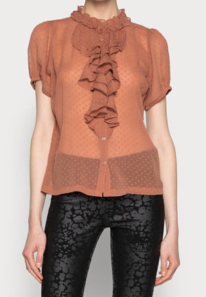 Przezroczysta bluzka w kolorze rdzy z teksturowanymi groszkami, falbanką z przodu, krótkimi bufiastymi rękawami, w połączeniu z czarnymi spodniami w wzory.