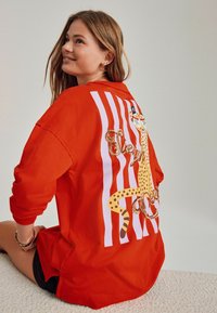 Cămașă roșie cu mânecă lungă, având un model cu dungi albe și un grafic cu leopard, decorat cu paiete pe spate. Croială lejeră și material moale.