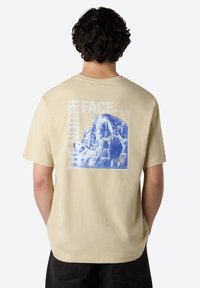 Béžové bavlněné tričko s modrou grafikou hory a textem "THE NORTH FACE". Krátké rukávy, volný střih a kulatý výstřih.