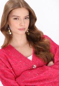 Helles pinkes Cardigan mit V-Ausschnitt, verziert mit mehrfarbigen Perlen und einem einzigen Knopfverschluss. Lange, wellige braune Haare.