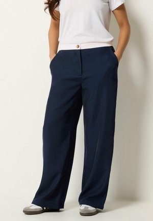 Femme portant un pantalon large bleu marine avec une ceinture blanche boutonnée, un t-shirt blanc rentré, les mains dans les poches, et des baskets blanches.