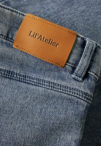 Jeans de mezclilla con un parche de cuero marrón claro con el texto grabado "Lil'Atelier", una tela azul texturizada y detalles cosidos en la cinturilla.