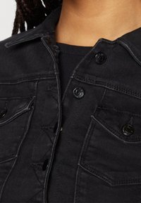 Černá džínová bunda s knoflíkovým zapínáním, špičatým límcem, dvěma náprsními kapsami a jemným texturovaným detailem na látce.