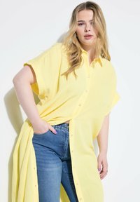 Camisa amarilla de botones con mangas cortas y una tela texturizada, combinada con jeans ajustados azules. La camisa está abierta por los lados.