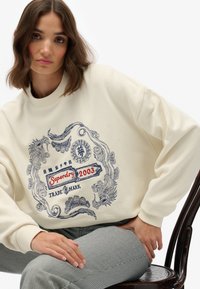 Crème sweatshirt met een textuurpatroon in marineblauw en rood, met de tekst "Superdry 2003" en verfijnde versieringen. Gestreepte broek zichtbaar.