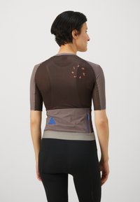 Short-sleeved athletic shirt in brown and gray fabric, featuring mesh accents, a printed logo, and a zippered pocket on the back.