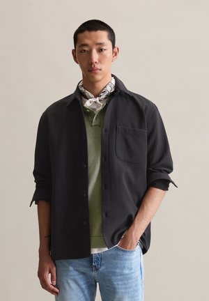 Black button-up shirt with a textured fabric, worn over a green polo. The model accessorizes with a patterned scarf and blue jeans.