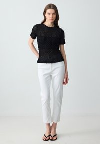 Top in maglia nera a texture, con maniche corte, scollatura rotonda e design a strati, abbinato a pantaloni bianchi a gamba dritta e infradito neri.
