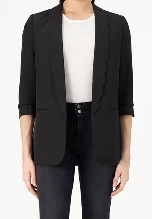 Frau trägt einen schwarzen Blazer mit wellenförmigen Revers, Dreiviertelärmeln, weißem T-Shirt und hoch taillierter schwarzer Jeans mit Doppelknöpfen.