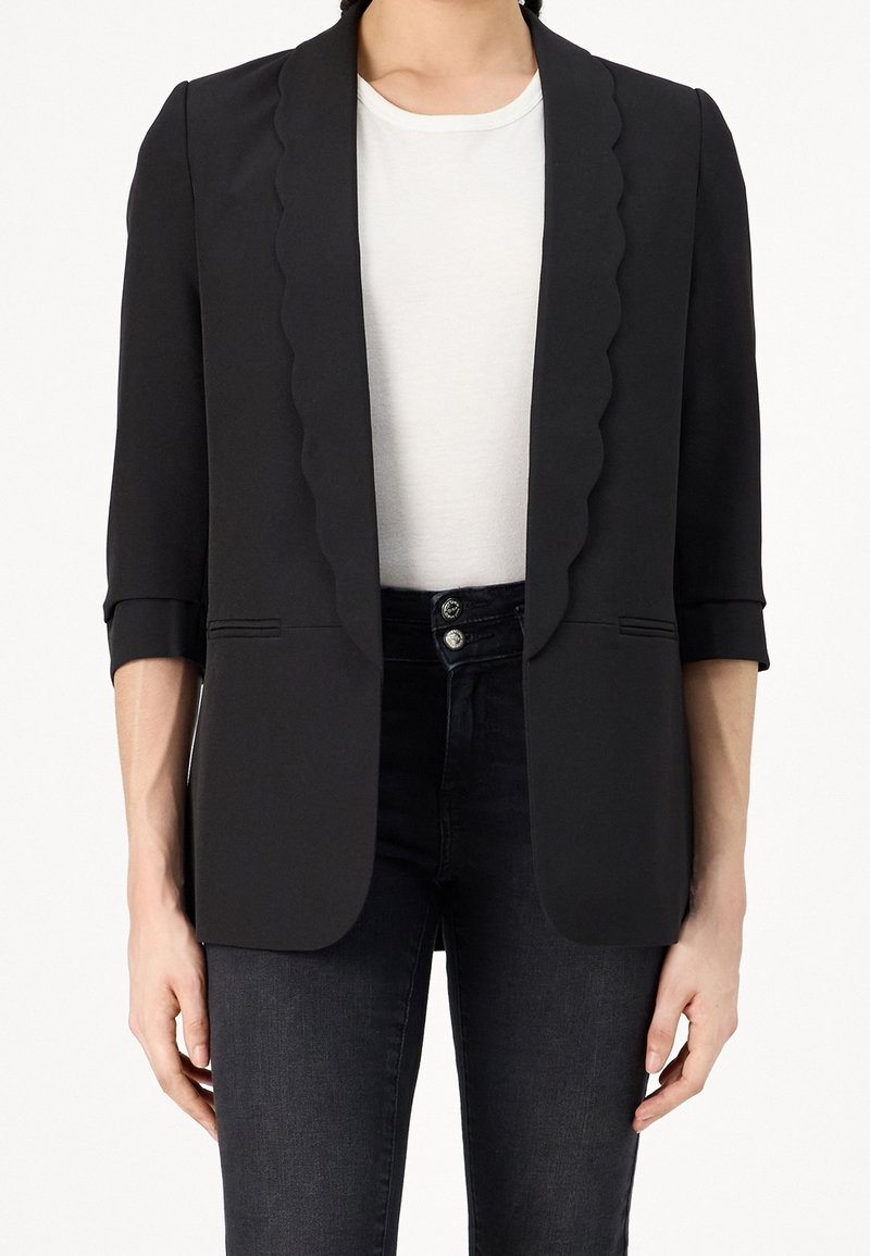Femme portant un blazer noir avec des revers festonnés, des manches trois-quarts, un t-shirt blanc et un jean noir taille haute avec double boutons.