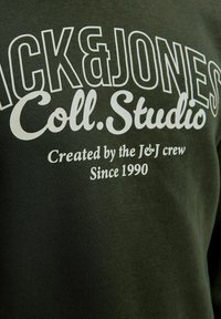 Ciemnozielona bluza z dużym białym napisem: "JACK&JONES Coll. Studio" oraz "Stworzona przez ekipę J&J od 1990 roku." Miękka struktura materiału.