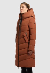 Brauner, gepolsterter Wintermantel mit hohem Kragen, Reißverschluss und Seitentaschen. Das Design weist diagonale Steppmuster und eine verstellbare Kapuze auf.